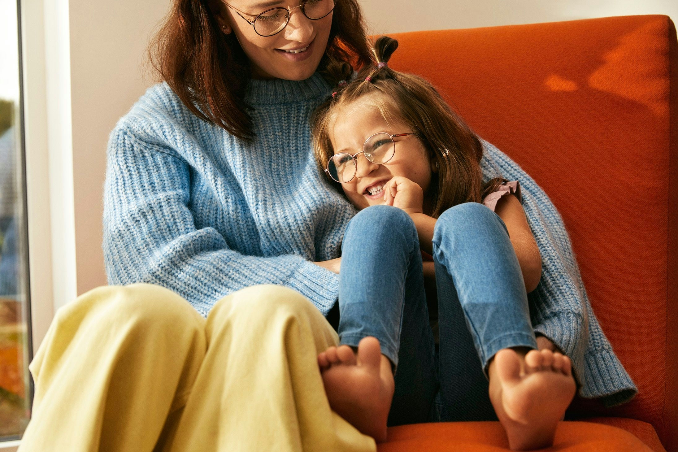 Mor sitter med datter armkroken i en oransje stol. Moren ser ned på datteren og datteren smiler og ser ut i rommet.