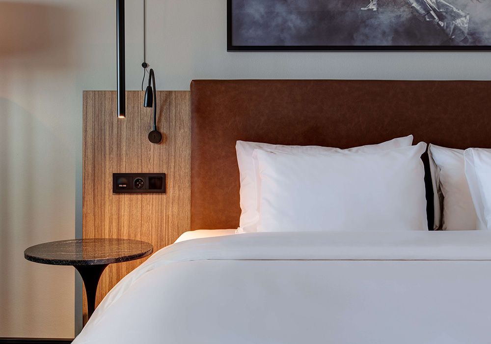 Seng på hotellrom med hvitt sengesett og brun gavl i skinn og eik.