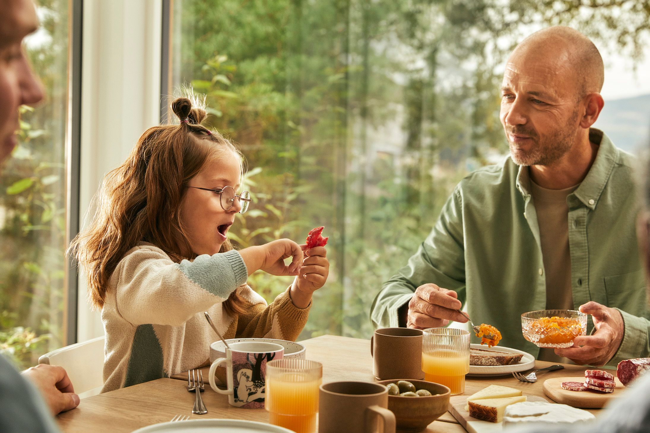 Ung jente og mann sitter rundt et frokostbord i grønne omgivelser.