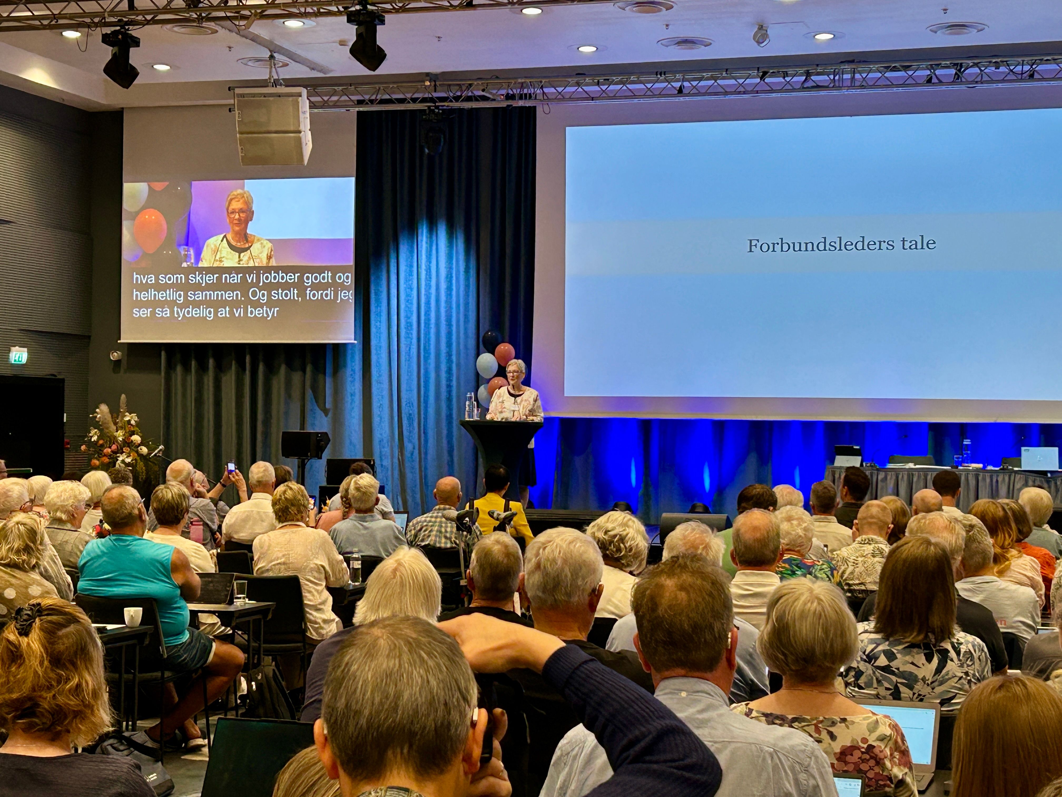En kvinne taler foran en større forsamling i en konferansesal.