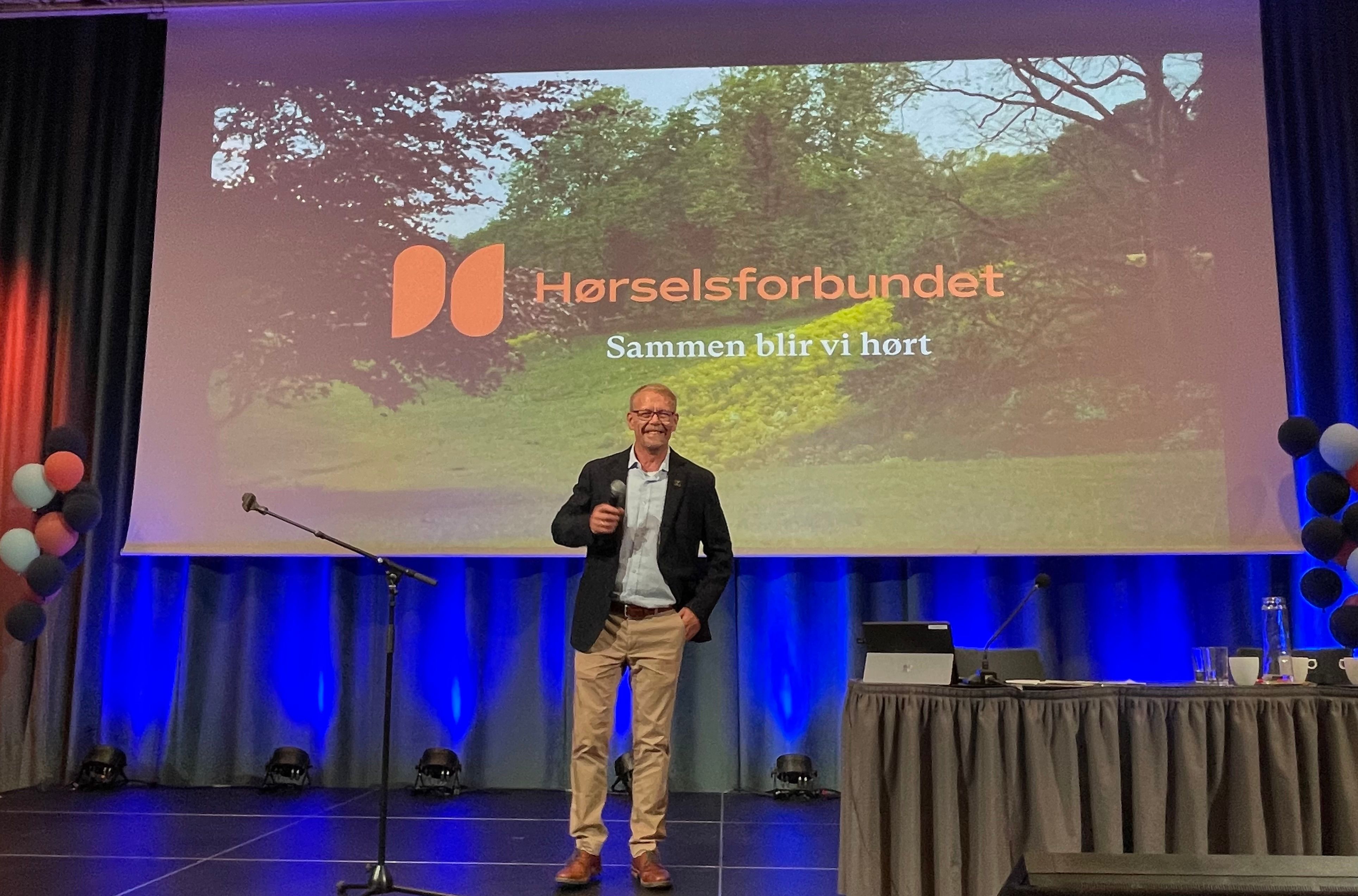 Smilende mann på en scene med teksten Hørselsforbundet i bakgrunnen
