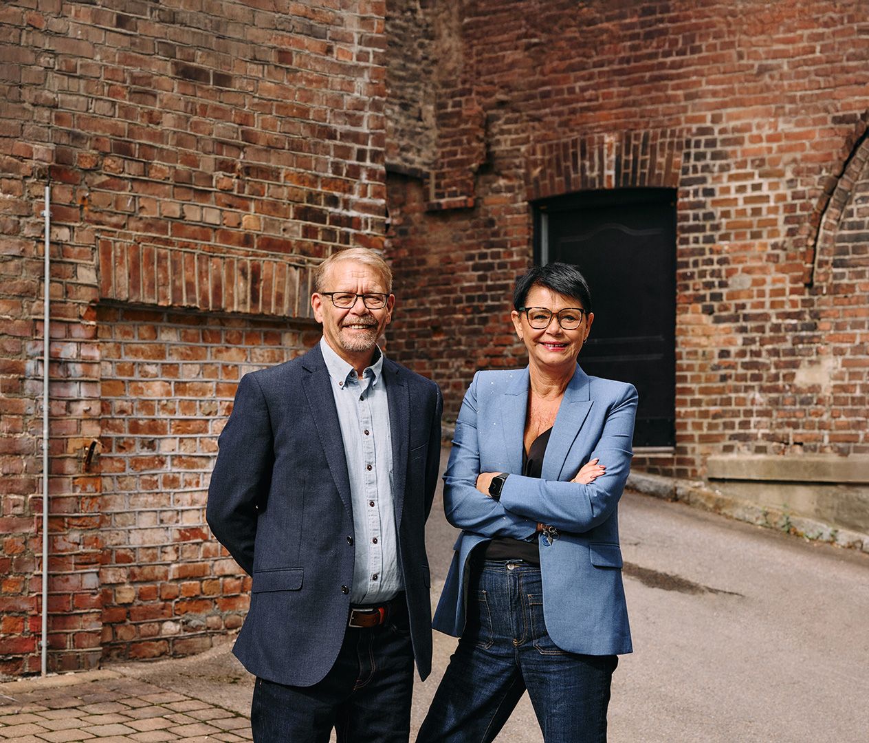 Forbundsleder Bjørn-Tore Lunde og generalsekretær Inger Helene Venås stående side om side, smilende og vendt mot kameraet med blå dressjakker og jeans, foran en rustrød murvegg