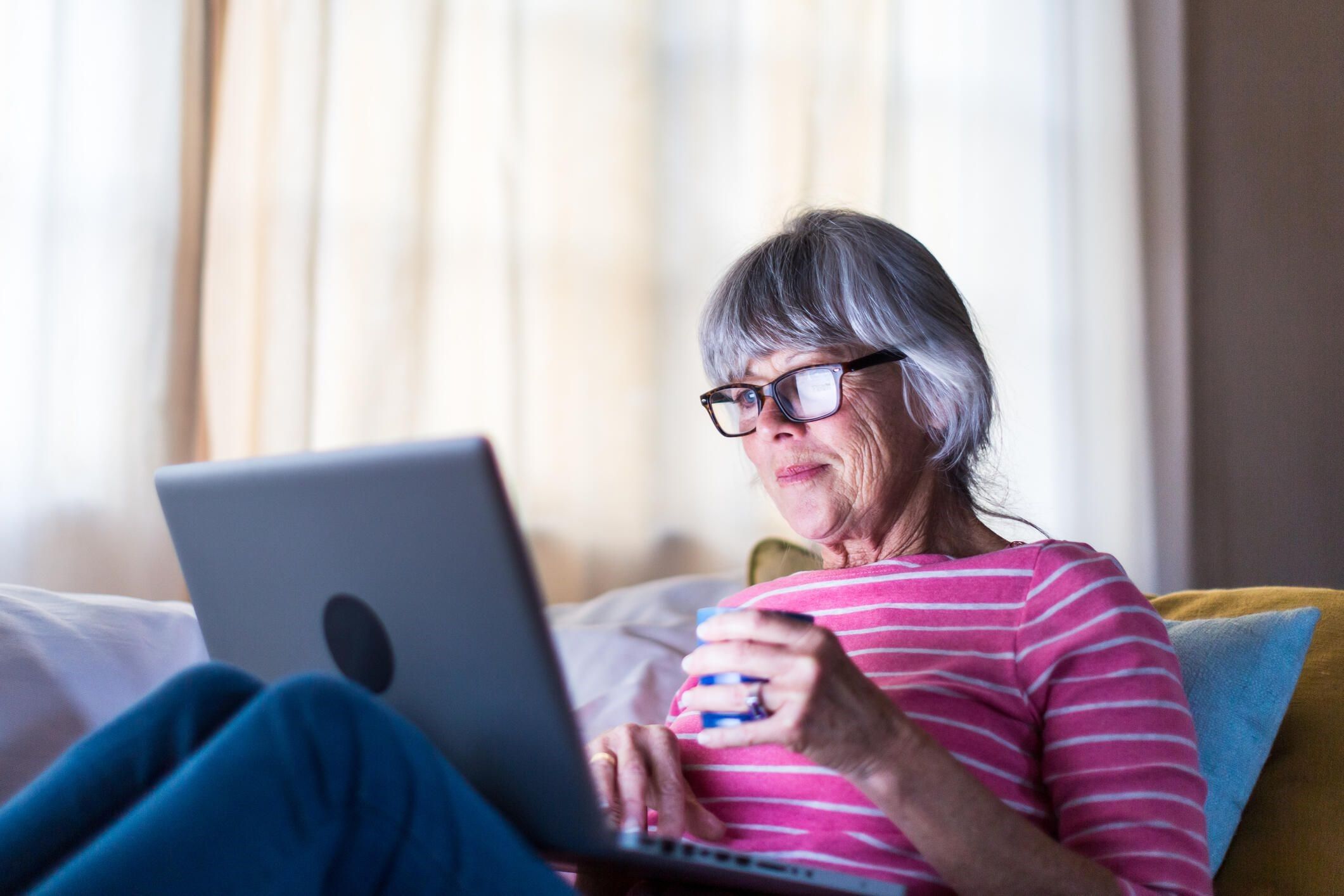 Voksen dame sitter behagelig i sofaen med en laptop på fanget og en kopp med varm drikke i hånden mens hun ser på skjermen.