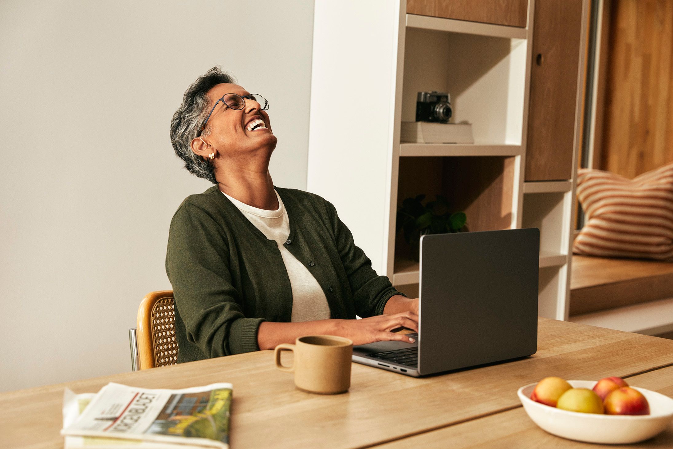 Kvinne sitter foran laptop ved bord, kaster hodet bakover og ler