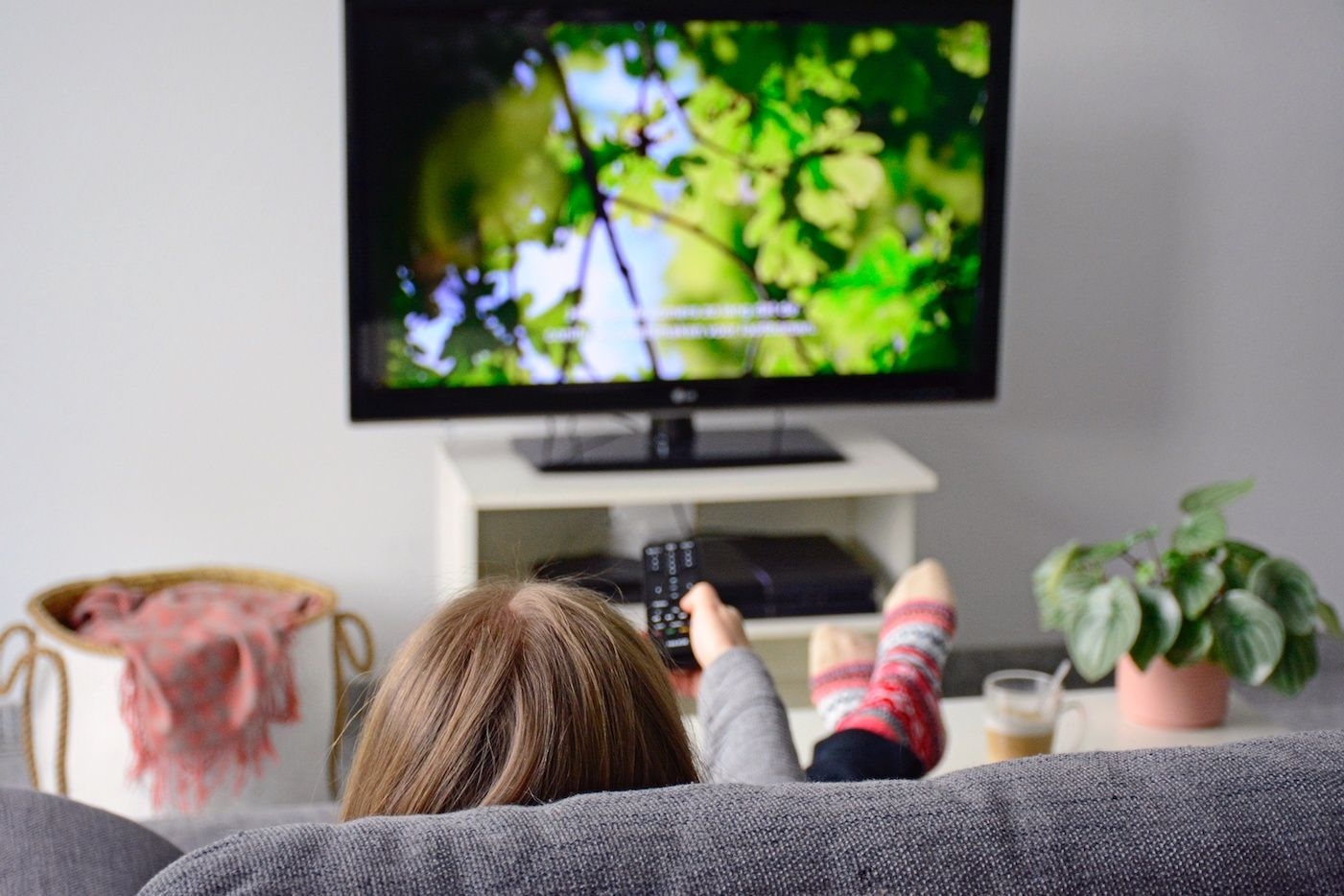 Kvinne i sofa foran TV med fjernkontroll i hånda og teksting på TVen