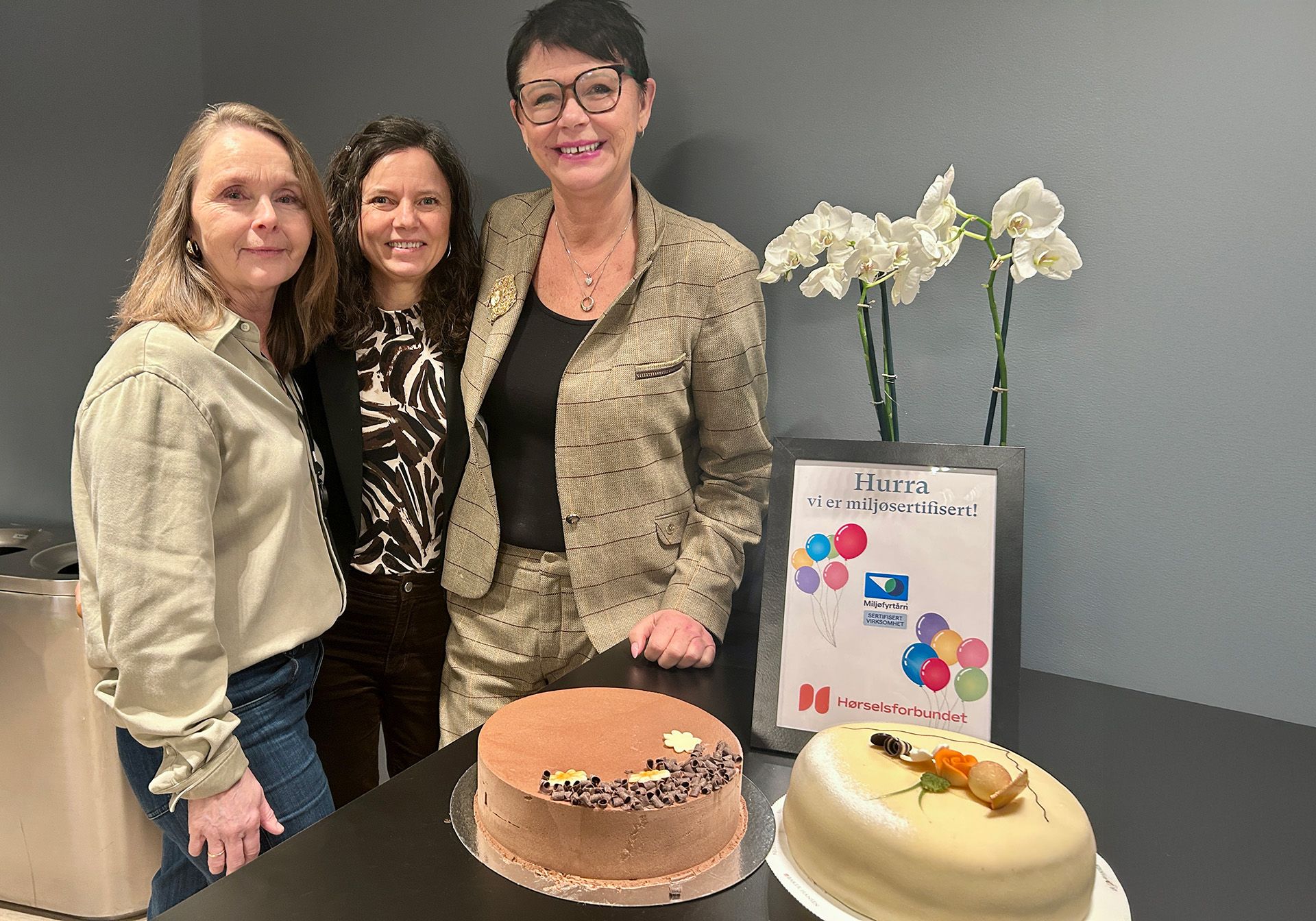 Tre kvinner smiler mot kamera, ved siden av et bord med to kaker på og en plakat med teksten Hurra vi er miljøsertifisert!
